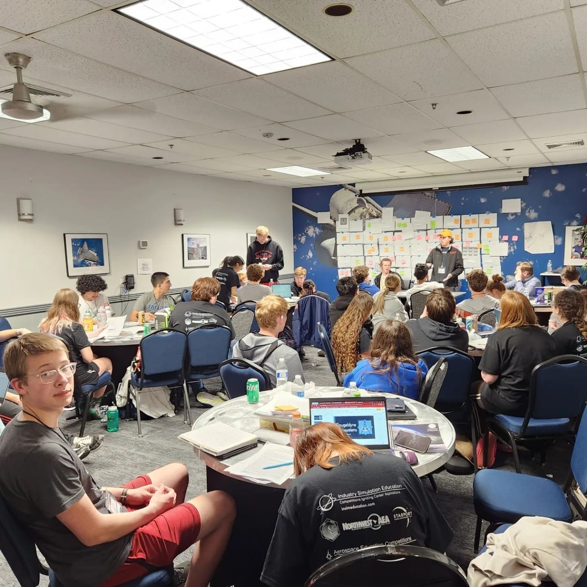 A competition mission room with students at tables in front of a wall of design notes